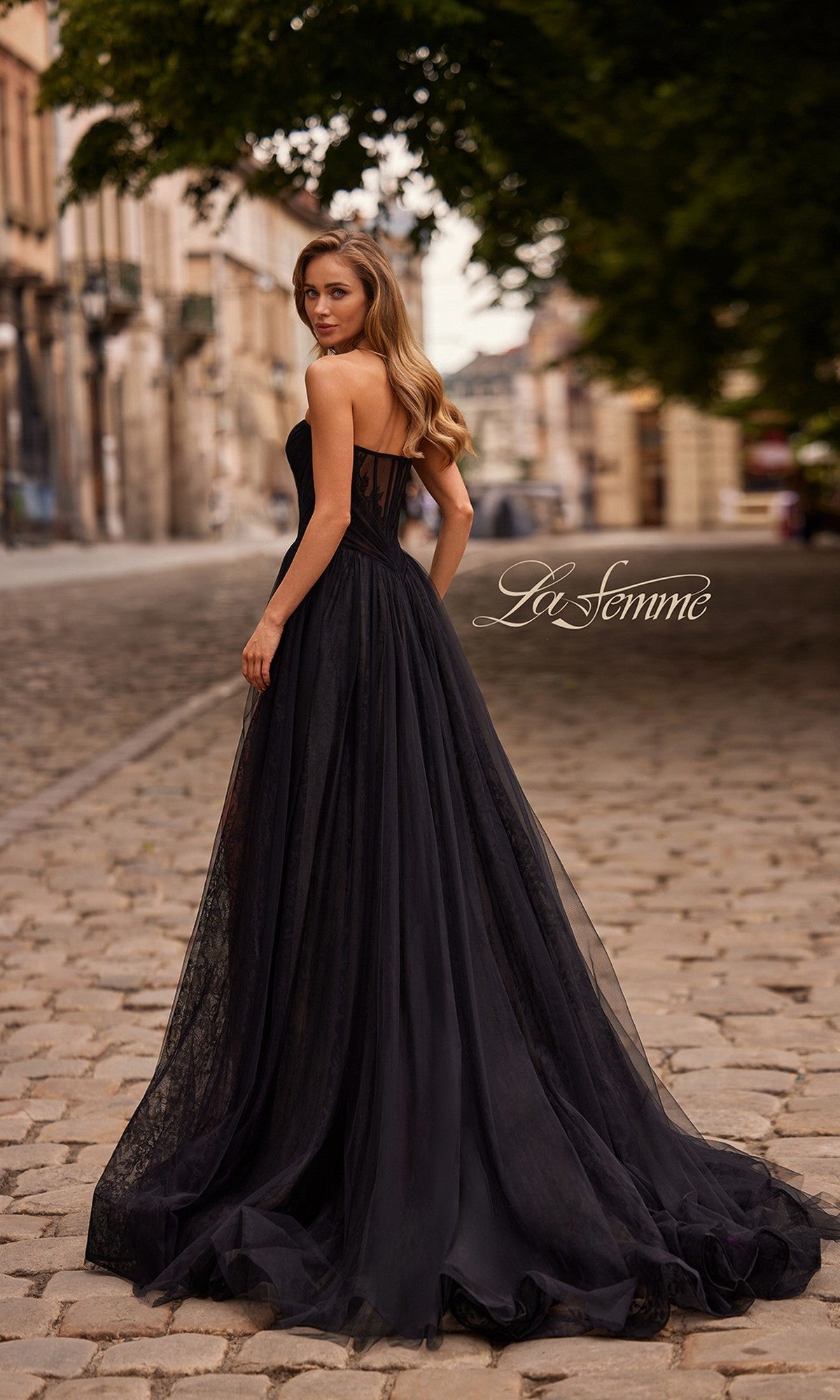 A woman wearing the Long Prom Dress: La Femme 33307 with a flowing train stands on a cobblestone street, looking over her shoulder. The words